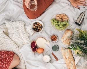 Weiße Picknickdecke mit Tellern, Schalen, Käse, Obst, Weißbrot, Weintrauben und Blumen