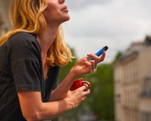 Eine Frau im T-Shirt steht  auf einem Balkon, raucht eine blaue Einmal-E-Zigarette und hält in der anderen Hand eine rote Tasse.