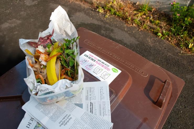 Bioabfälle wie Bananenschalen und Gemüseabschnitte liegen eingewickelt in Zeitungspapier auf einer braunen Biotonne. Auf der Tonne klebt ein Hinweisaufkleber gegen Plastiktüten. In Zeitungspapier eingewickelte Bioabfälle liegen auf einer Biotonne.