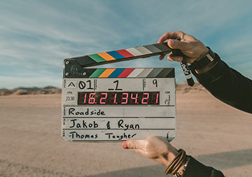 Eine Filmklappe wird von einer Hand gehalten; im Hintergrund blauer Himmel und Sandboden.
