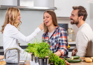 Eine Familie steht in der Küche; das Mädchen sitzt auf der Küchenzeile und reicht