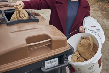 Eine Frau wirft gefüllte Papierbeutel aus dem Bringsystem-Eimer in einen Bioabfallccontainer