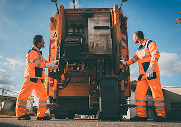 Zwei Müllwerker stehen am Müllfahrzeug; die Tonne wird gekippt.