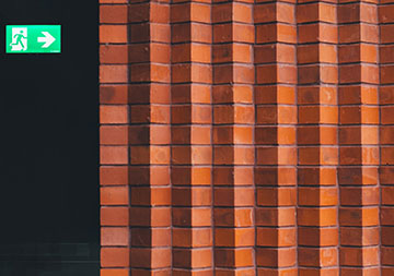 Ein beleuchtetes Notausgang-Schild vor schwarzem Hintergrund links; rote Backsteinmauer rechts